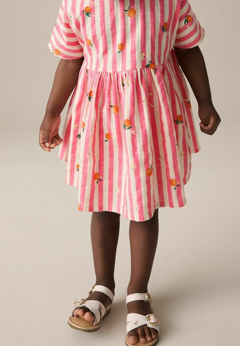 Enfant portant une robe rayée rose et blanche avec un imprimé fruit orange et des sandales blanches, debout sur un fond neutre.