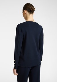 Donna con i capelli raccolti in un chignon, che indossa un maglione blu navy con bottoni dorati sulle maniche, abbinato a pantaloni blu navy, di spalle su uno sfondo bianco.