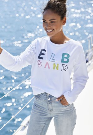 White long-sleeve sweatshirt with multicolored letters spelling "ELB SAND," paired with light blue high-waisted jeans, on a boat by the water.
