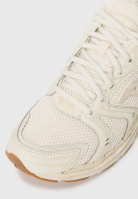 Scarpa sportiva beige con tomaia in rete, dettagli lisci e suola in gomma. Presenta lacci e un design a punta rotonda.