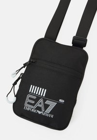Borsa a tracolla nera in nylon con chiusura a zip, caratterizzata da un logo bianco e un design a strisce. Tracolla regolabile e superficie testurizzata per una migliore impugnatura.