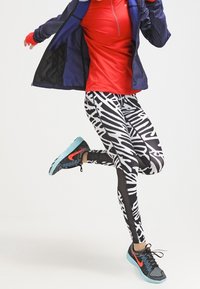 Maglietta rossa con zip e maniche lunghe, giacca blu navy, leggings a stampa zebra bianca e nera, e scarpe sportive nere con accenti turchesi.
