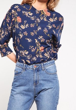 Kobieta w granatowej, kwiecistej bluzce na guziki z rękawami do łokci, włożonej w jasnoniebieskie, wysokiej talii jeansy.