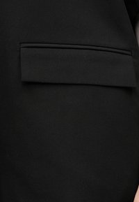 Tissu noir avec un détail de poche à rabat horizontale, légèrement surélevée par rapport à la surface, sur un vêtement en gros plan.