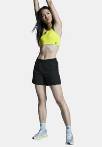 Geel sportbeha, zwarte shorts, grijze sneakers met gele accenten. Model staat met opgetrokken armen en toont een strakke atletische uitstraling.
