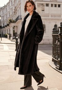 Palton din blană artificială neagră, cu un guler larg, purtat deschis; asociat cu pantaloni negri cu talie înaltă și pantofi cu vârful ascuțit, pe o stradă din oraș.