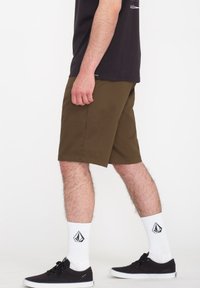 Braune Shorts aus leichtem Material, mit einem einfachen Saum, kombiniert mit schwarzen Sneakers und weißen Socken mit einem Logo.