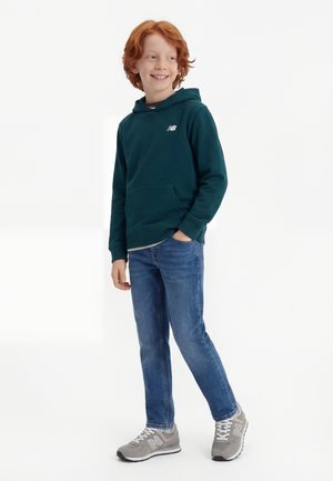 Ragazzo dai capelli rossi sorridente, che indossa una felpa con cappuccio verde scuro, jeans blu, sneakers grigie, in piedi con una mano in tasca su sfondo bianco.