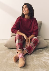 Punane dressipluus, ruudulised kodupüksid punase, valge ja rohelise aktsendiga ning karulooma motiiviga sulejoped sokid. Istub padjal, neutraalse taustaga.