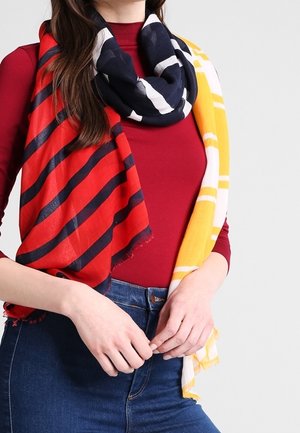 Femme portant un haut à manches longues rouge et un jean bleu, avec une écharpe multicolore aux rayures rouges, bleu marine, jaunes et blanches.