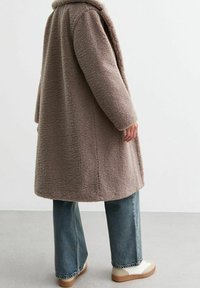 Donkerbruine, pluizige jas met een gestructureerd oppervlak, voorzien van een sjawl-kragen, lange mouwen en een losse pasvorm, gecombineerd met lichtblauwe flared jeans.