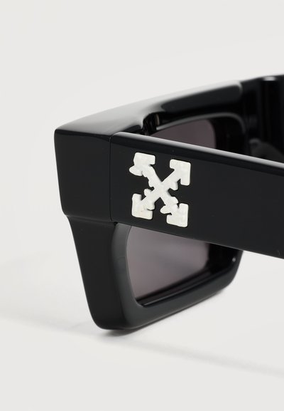 Lunettes de soleil rectangulaires noires avec une monture épaisse et un logo flèche croisée blanc sur la branche.
