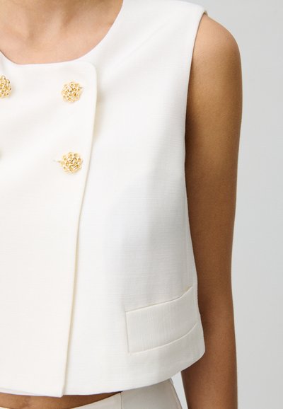 Top court sans manches blanc avec un tissu texturé, des accents floraux dorés et un petit détail de poche à l'ourlet.