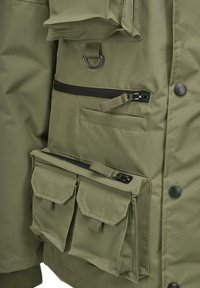 Chaqueta verde oliva con múltiples bolsillos, cremalleras y cierres a presión. La tela tiene una textura suave con elementos de diseño centrados en la utilidad.