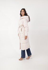 Abrigo acolchado de color rosa claro con capucha, cinturón en la cintura y diseño de cuadros. Llevado sobre pantalones de mezclilla oscuros y sandalias de tacón transparente.