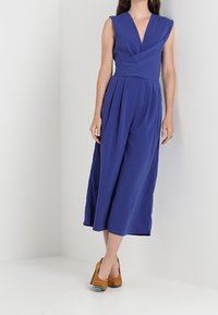 Combinaison sans manches à décolleté en V profond de couleur bleu roi uni, avec jambes larges et plis. Le tissu lisse tombe élégamment, associé à des talons marron.