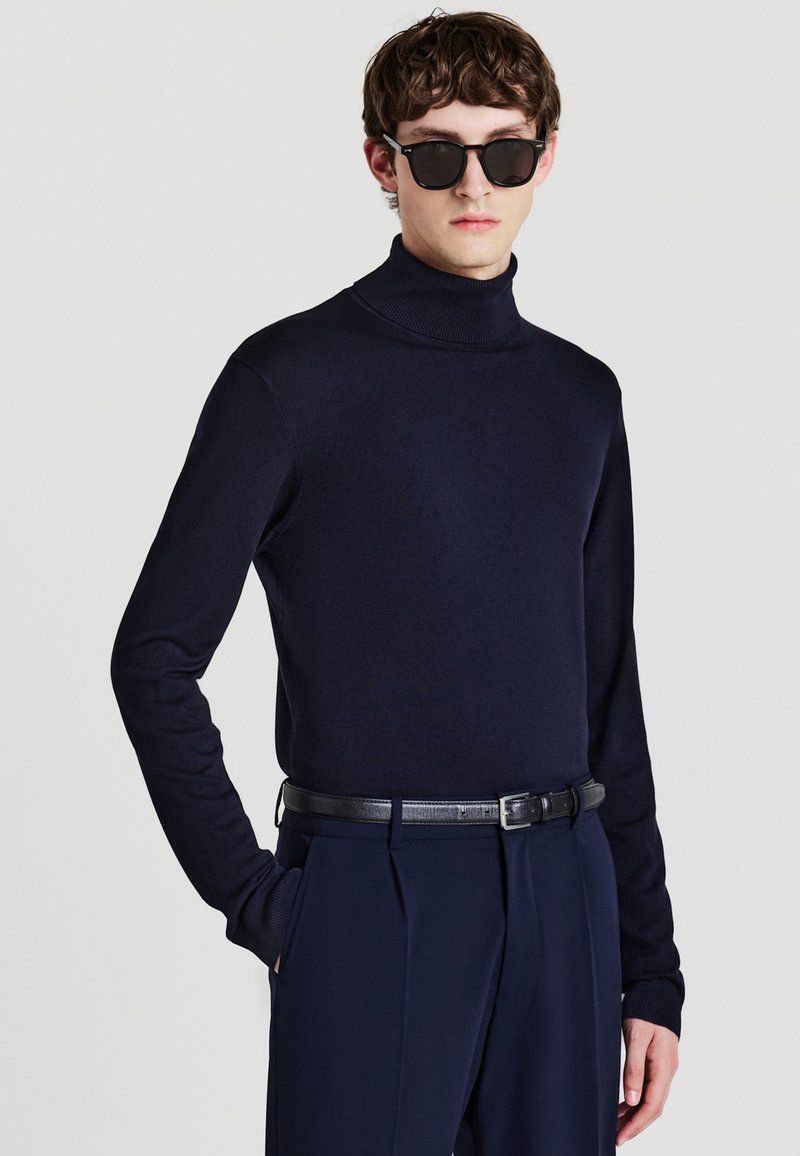 Navyfarbener Rollkragenpullover aus Strickstoff, kombiniert mit maßgeschneiderten navyfarbenen Hosen und einem schwarzen Ledergürtel, getragen mit Sonnenbrille.