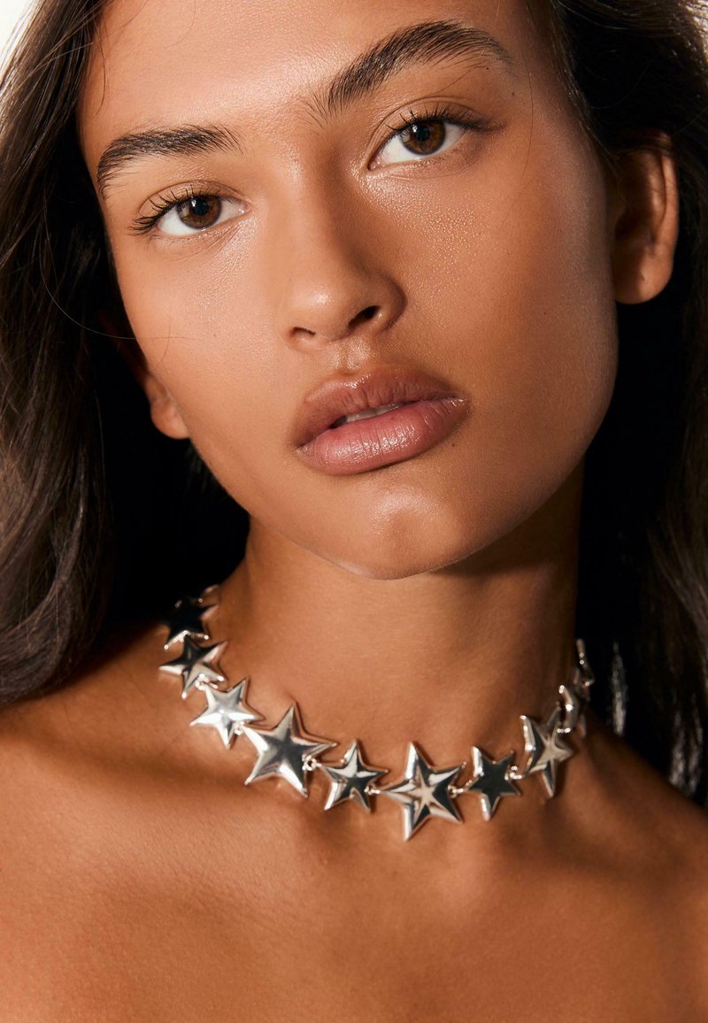 Sølvfarvet stjernesmykke i choker-stil med en skinnende, metallisk finish. Har sammenkoblede stjerner i forskellige størrelser, der skaber et dristigt design.