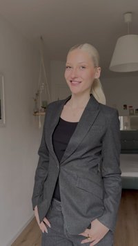 Szürke, testhezálló szabású, szabott blézer, melynek jellegzetes kivágású nyakkendői és két elülső zsebe van, fekete felsővel viselve. Sima textúra, minimalista dizájn.