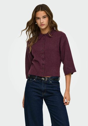 Jeune femme portant un pull prune boutonné avec des manches trois-quarts et un jean bleu foncé avec une ceinture noire.