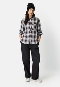 Kariertes Hemd in Schwarz und Grau, langärmlig, Ärmel bis zu den Ellbogen hochgekrempelt, mit Brusttasche; kombiniert mit weit geschnittenen schwarzen Hosen und einer schwarzen Beanie-Mütze.