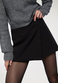 Mini gonna nera, sagomata, con davanti piatto e tasca laterale. Abbinata a un maglione grigio a maglia e collant neri trasparenti.