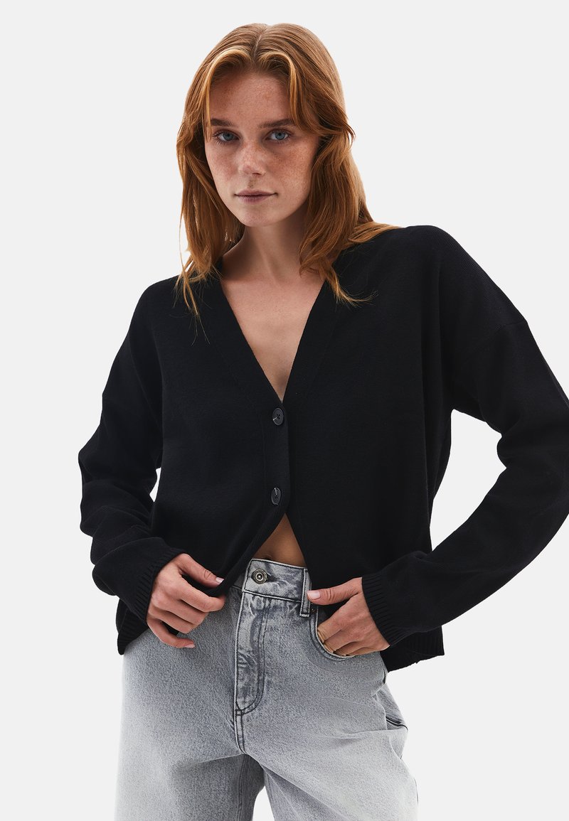 Mustat neulotut neuletakit, joissa on syvä V-pääntie ja kaksi etupainiketta. Varustettu ribbadatuilla hihansuilla ja helmalla. Puettu vaaleanharmaiden korkeavyötäröisten farkkujen päälle.