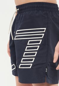 Marina simbyxor med en vit geometrisk design på sidan, med resårmidja, dragsnöre och sidsplittar för enkel rörelse.