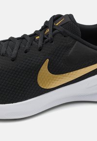 Crni atletski tenisice s gornjim dijelom od teksturirane tkanine, zlatnim Nike swoosh logom i bijelim gumenenim potplatom. Torbice i jezik su u odgovarajućoj crnoj boji.
