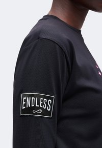 Gros plan d'une personne portant une chemise noire avec un écusson rectangulaire sur la manche affichant "ENDLESS" et un symbole infini en dessous.