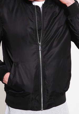 Personne portant une veste bomber noire zippée avec poignets et ourlet côtelés, une main dans la poche, sur un fond clair uni.