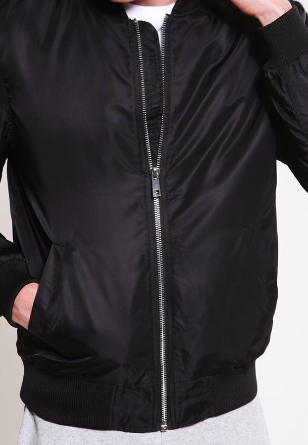 Personne portant une veste bomber noire zippée avec poignets et ourlet côtelés, une main dans la poche, sur un fond clair uni.
