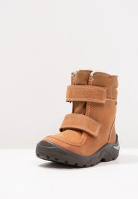 Brązowy skórzany but z miękkim futerkiem, dwiema regulowanymi paskami na rzep i wytrzymałą czarną gumową podeszwą zapewniającą przyczepność.