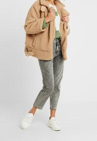 Chaqueta beige, esponjosa y de gran tamaño con cuello y correas laterales, llevada sobre un suéter de rayas verdes y blancas, combinada con jeans grises y zapatillas blancas.