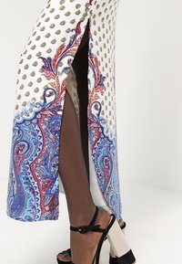 Robe maxi en tissu blanc avec un motif paisley bleu et rouge, dotée d'une fente sur le côté. Talons noirs avec des semelles épaisses et des brides à la cheville.