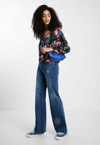 Florale Bluse mit langen Ärmeln in Schwarz, kombiniert mit blauen Schlagjeans mit bestickten Blumenmustern und blauen Turnschuhen.