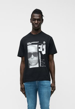 Mann trägt ein schwarzes T-Shirt mit Karl Lagerfeld Jeans Schriftzug, Gesichtsprint und Kamera-Illustration, kombiniert mit blauen Jeans, steht vor weißem Hintergrund.