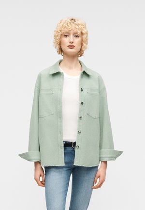 Woman with short curly blonde hair wearing a light green corduroy shirt jacket, white T-shirt, and blue jeans against a plain background.