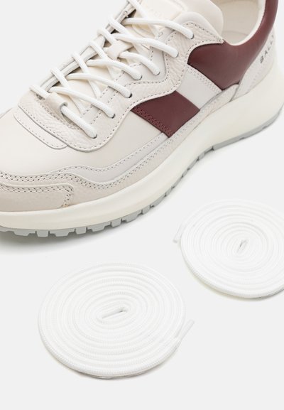 Κοντινό πλάνο ενός μπεζ και μπορντό αθλητικού παπουτσιού με λευκές κορδόνες, δίπλα σε δύο τυλιγμένα επιπλέον λευκά κορδόνια σε μια λευκή επιφάνεια.