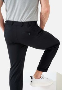 Schwarze Hose mit einer taillierten Passform, elastischem Bund und zwei Gesäßtaschen mit Knöpfen. Kombiniert mit weißen Sneakers.
