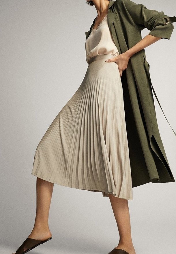Femme portant une jupe midi plissée beige, un top en satin crème, un long manteau vert olive et des chaussures marron sans lacets, posant avec une main sur la hanche.