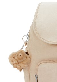 Zaino in tessuto beige con patta pieghevole, tasca frontale con zip e un portachiavi a forma di scimmia in peluche attaccato da un anello metallico.