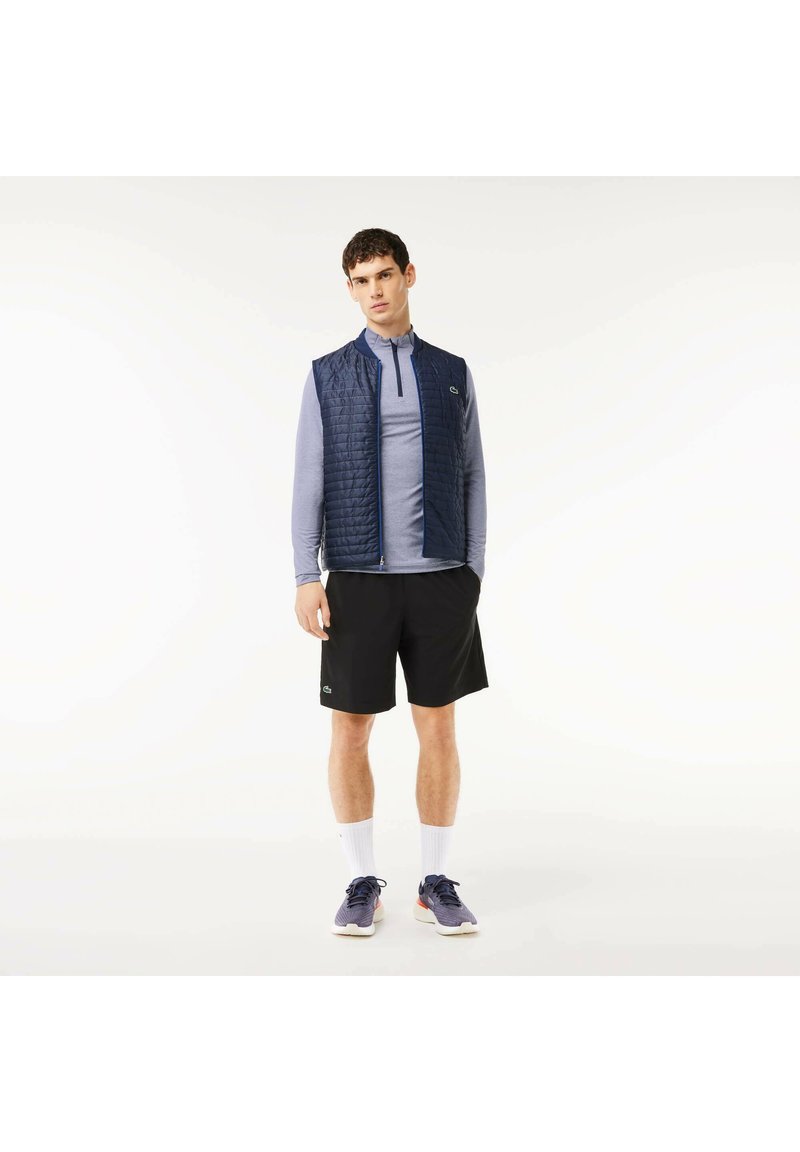 Männernavy gesteppte Weste über einem hellblauen Langarmhemd, schwarzen Shorts, weißen Socken und blauen Sportschuhen. Einfaches Design und Materialien.