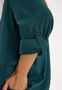 Chemisier bleu sarcelle avec des manches retroussées, présentant des détails de poignets boutonnés. Texture de tissu lisse, avec une coupe ample et une silhouette droite.