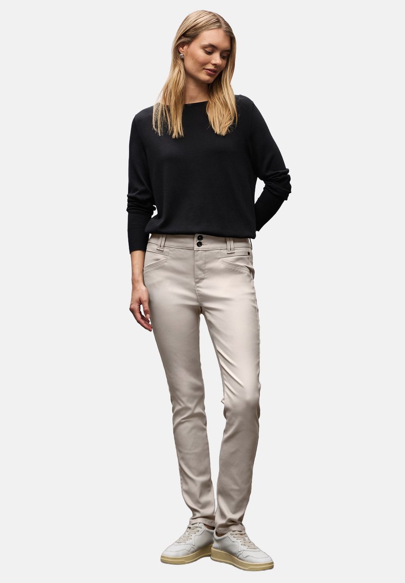 Světlé béžové skinny kalhoty s dvěma předními kapsami, dvouknoflíkovým zapínáním a hladkým povrchem. S kombinací s černým svetrem s dlouhým rukávem a bílými teniskami.