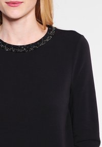 Černý top s dlouhým rukávem a korálkovým límcem. Hladká látka, přiléhavý střih, kulatý výstřih a jemný lesk zdobení.