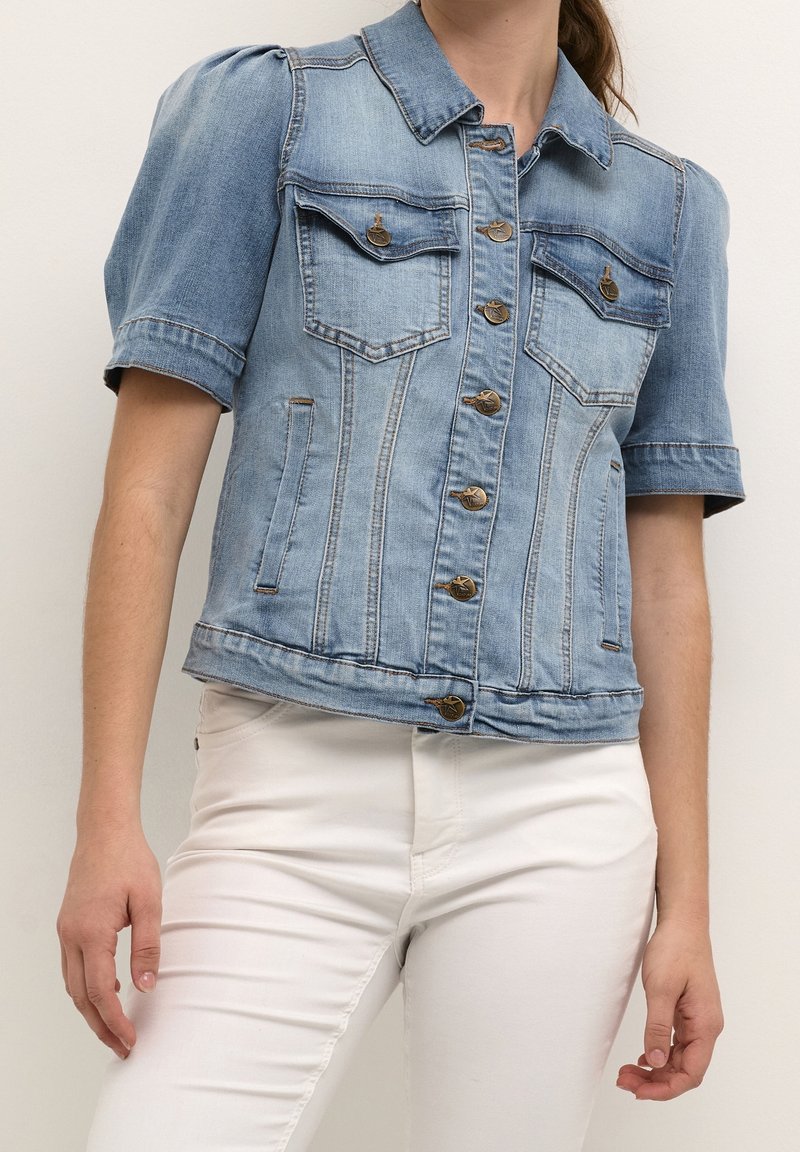 Giacca di jeans azzurro chiaro con maniche corte a sbuffo, tasche frontali e chiusure a bottoni in bronzo. Presenta una forma aderente e dettagli cuciti.