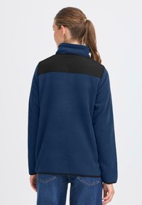 Giacca in pile di colore blu navy con una spalla nera. Presenta un colletto alto, maniche lunghe, una texture liscia e una vestibilità comoda.