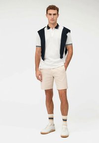 Weißes Polohemd mit dunklen Akzenten, beige Shorts, weiße Turnschuhe und gestreifte Socken. Ein marineblauer Pullover ist über die Schultern gelegt.