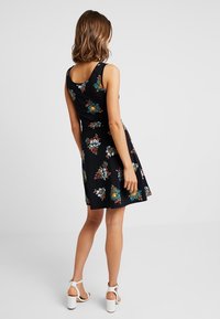 Černé bezrámové šaty s květinovým vzorem v tyrkysové, oranžové a bílé barvě. Silueta do A s hladkým povrchem. Kombinováno s bílými sandály na blokovém podpatku.